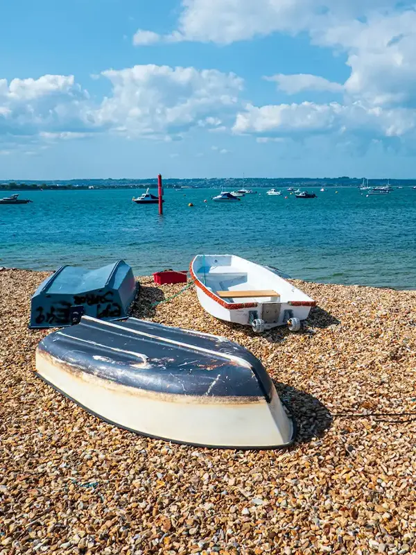 Petits bateaux sur une plage de galets avec voiliers ancrés au large sous un ciel bleu.