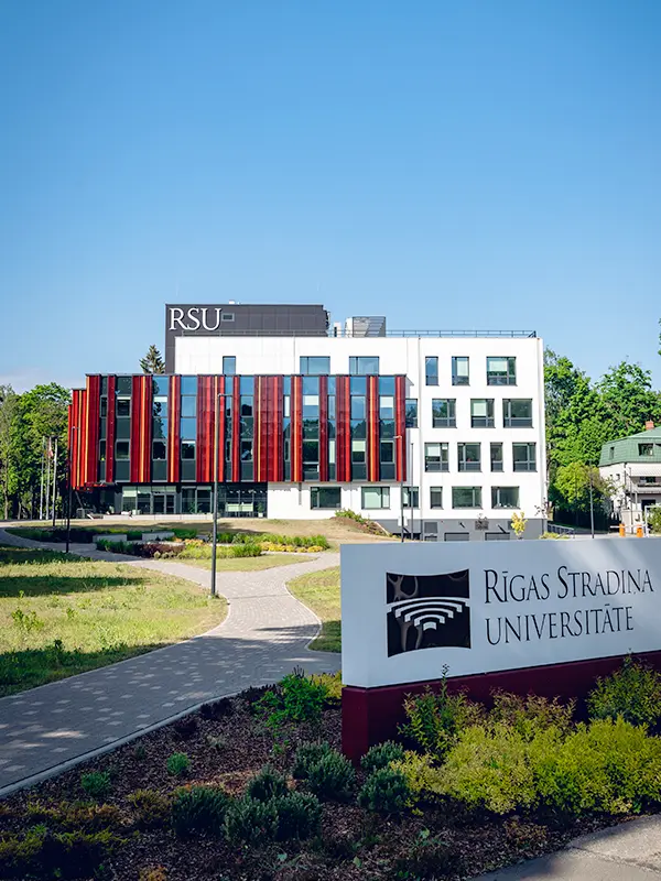 Bâtiment moderne blanc de l'RSU avec panneaux verticaux rouges et panneau Rīgas Stradiņa Universitāte entouré de jardins verdoyants par beau temps